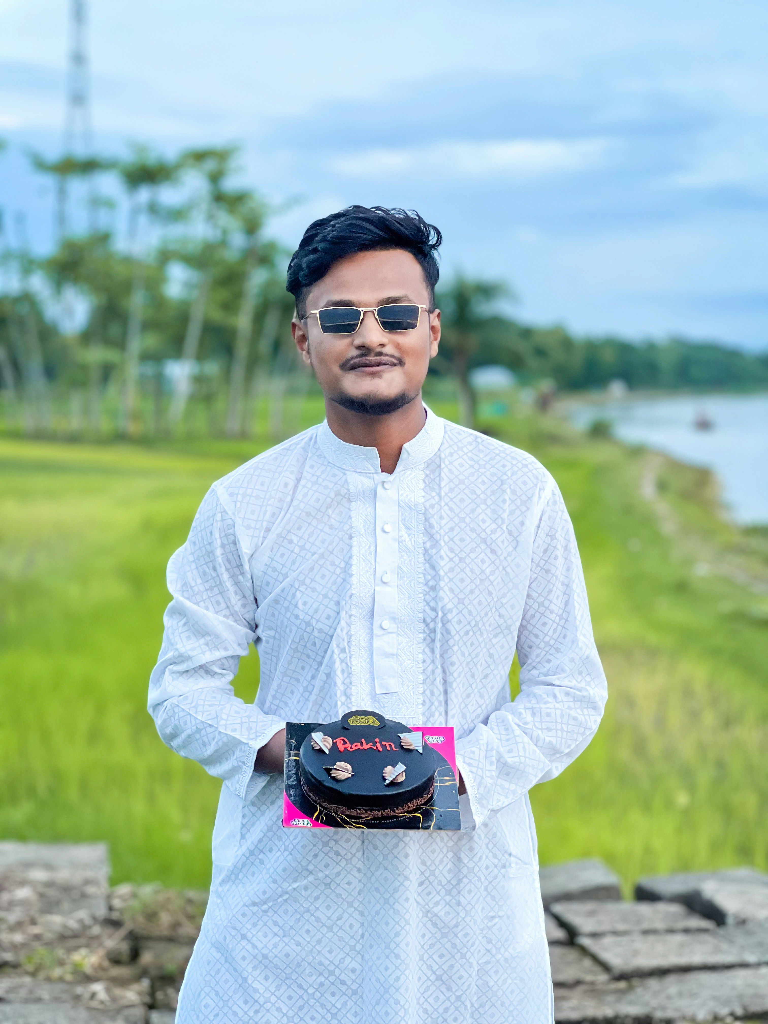 A man in a white patterned kurta holding a small chocolate cake outdoors